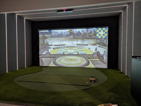 Indoor golf simulator setup with large projection screen displaying a golf course and putting green mat on artificial turf floor.