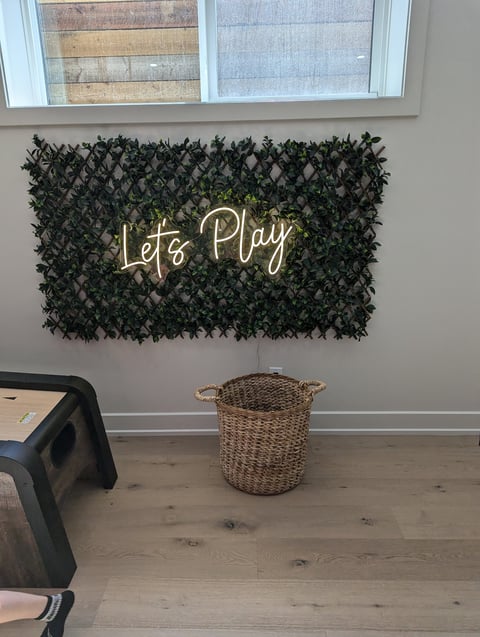 Gold neon Lets Play sign mounted on a dark green leaf wall panel above a woven basket in a playroom
