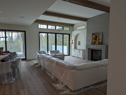 Modern living room under renovation with plastic-covered furniture, wood beams, skylights, and large windows overlooking outdoor scenery