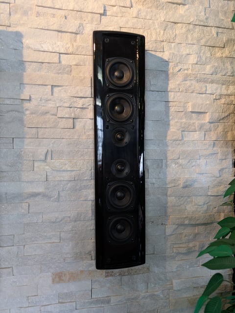 Black wall-mounted speaker with six drivers mounted on a light brick wall