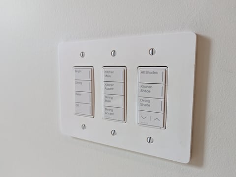 Wall-mounted lighting control panel with three vertical menu options for Rooms, Kitchen, and All Shades, mounted on white wall