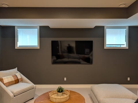 Modern living room with taupe walls, two windows, wall-mounted TV, and light-colored seating with a round wooden side table