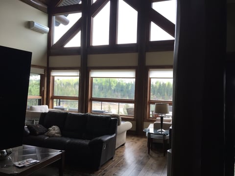 Modern living room with floor-to-ceiling windows overlooking forested mountains, black leather furniture, and wooden flooring