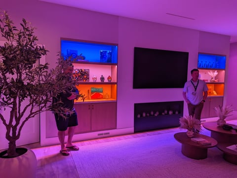 Modern living room with vibrant pink and blue LED lighting, wall-mounted shelves, fireplace, flat-screen TV, and two people viewing the display.